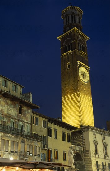 ITALY, Veneto, Verona, "Torre dei Lamberti clock tower in the Piazza Erbe illuminated at night part seen behind faded building facades, statue and lights and umbrellas of cafe bar."