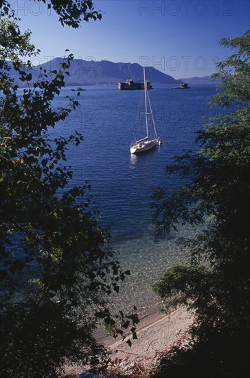 ITALY, Lake Maggiore, "Yacht anchored on lake framed by trees in foreground.  Stretch of pebble shore and clear, shallow water below."