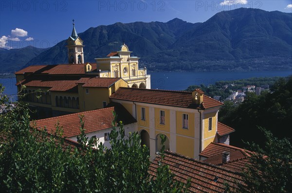 SWITZERLAND, Ticino, Lake Maggiore, Locarno.  Tiled rooftops and bell tower of yellow and white painted Church of Madonna del Sasso with lake and mountains beyond.