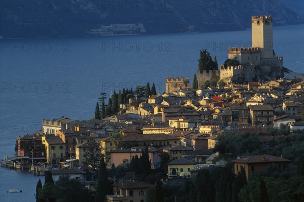 ITALY, Veneto, Lake Garda, "Malcesine.  View across town rooftops towards Castello Scaligero in warm, golden light with lake beyond."
