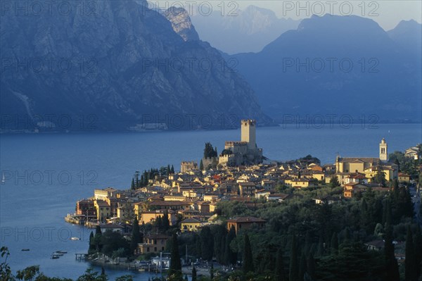 ITALY, Veneto, Lake Garda, "Malcesine.  View across town rooftops towards Castello Scaligero in warm, golden light with lake and mountains beyond."
