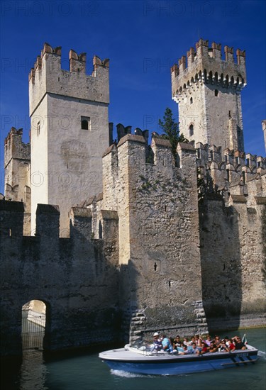 ITALY, Lombardy, Lake Garda , Sirmione.  Part view of crenellated walls and towers of the Rocca Scaligera medieval castle with passing tourist boat in foreground.