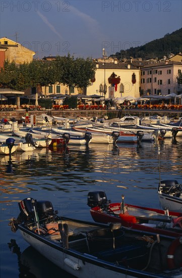 ITALY, Veneto, Lake Garda , Garda.  Harbour view with cafe tables set out on quayside overlooked by pastel coloured buildings and with moored boats in the foreground.