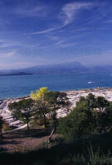 ITALY, Lombardy, Lake Garda , Sirmione.  View across Lake Garda from Villa Romana.  Flat outcrop of rock and trees in foreground with people sunbathing.  Distant mountains beyond.