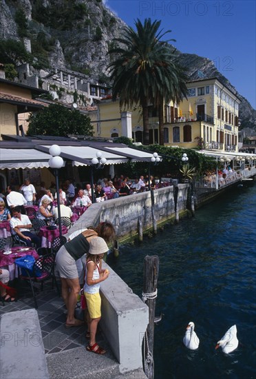 ITALY, Lombardy, Lake Garda , Limone Sur Garda.  Busy lakeside cafe with people sitting at tables under awning.  Woman and child feeding swans in the foreground.