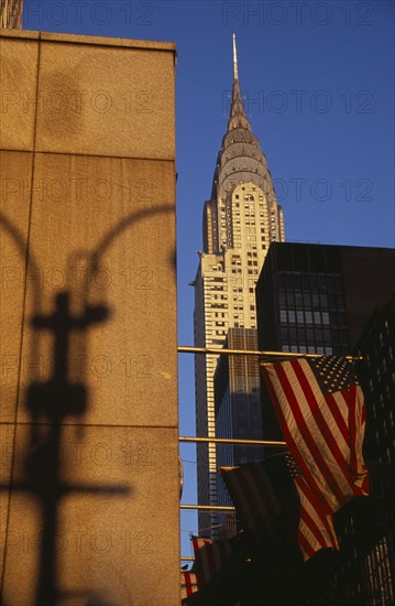 USA, New York, New York City, Part view of the Chrysler Building from East 42nd Street.  Seen above tower blocks and line of Stars and Stripes flags with shadow of street lamp on wall in foreground. Steel framed Art Deco skyscraper built 1928-1930 and designed by architect William Van Alen.