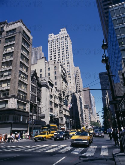 USA, New York, View along 5th Avenue in-between tall buildings with yellow taxi cabs on road and people walking on side walk