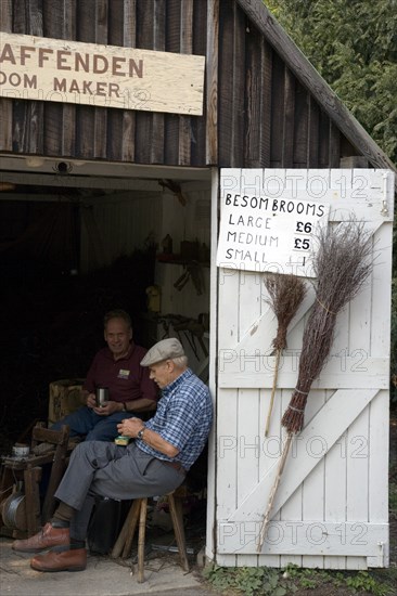 ENGLAND, West Sussex, Amberley, Amberley Working Museum. The Broom maker workshop with two men sitting in doorway next to display of brooms