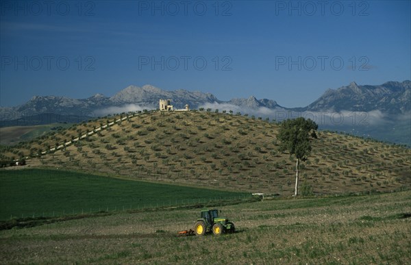 SPAIN, Andalucia, A tractor ploughing a terrace overlooked by a stone building on a hill and mountains with mist in the distance
