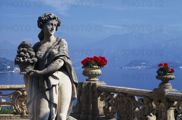 ITALY, Lombardy, Lake Como, Villa Balbianello.  Statue of woman holding basket of fruit with red geraniums in stone vessels on stone balustrade behind and view over lake.