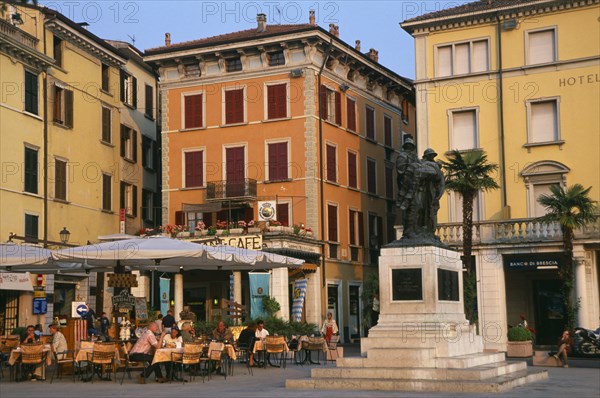 ITALY, Lombardy, Salo, "People sitting at outside cafe tables in piazza of town beside Lake Garda. Facades of buildings, bank and hotel painted pale yellow and orange with window shutters."