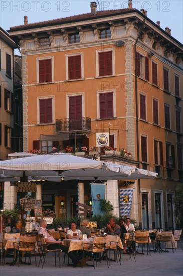 ITALY, Lombardy, Salo, People sitting beneath white awning at outside tables of cafe in piazza of town beside Lake Garda.   Facade of building behind painted orange with red window shutters.