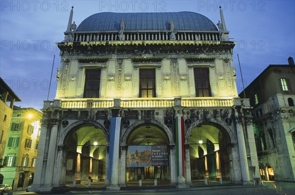 ITALY, Lombardy, Brescia, "Piazza della Loggia.  Facade of Loggia or town hall with arched entrance and colonnades, balcony and statues illuminated at night.  With poster advertising Monet exhibition"