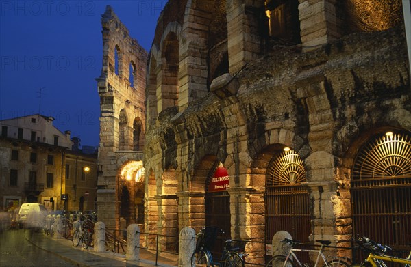 ITALY, Veneto, Verona, Piazza Bra.  Arena Roman amphitheatre completed in AD 30.  Part view of exterior at night with bicycles chained against railings in foreground and blur of passing traffic.