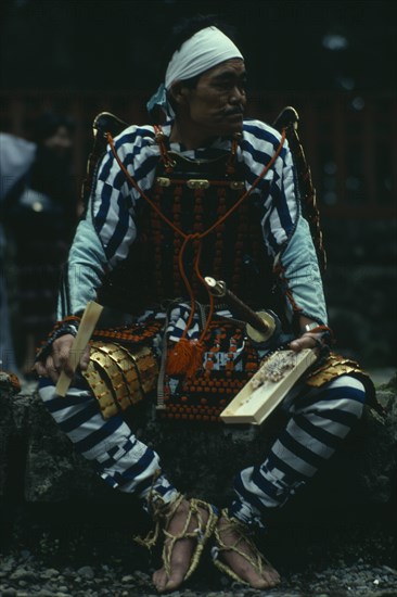 JAPAN, People, Samurai, "Portrait of Samurai wearing armour constructed of plates of metal or bamboo held together with coloured lacing, rope sandals and white cloth head band with sword at side.  Seated, holding bento lunchbox and chopsticks.  "