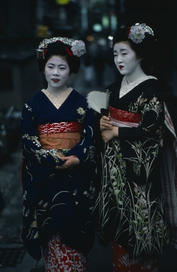 JAPAN, Customs, Geisha, Two maiko or apprentice geisha with white powdered faces and red painted lips wearing richly embroidered kimonos and decorative hair pieces.