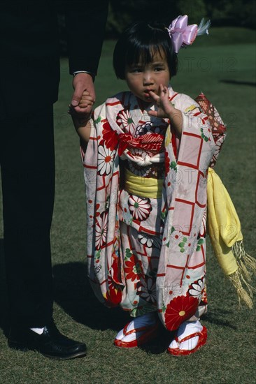 JAPAN, Honshu, Tokyo, Little girl holding the hand of a part seen guest at Shinto wedding ceremony wearing traditional dress printed with red and white squares and flowers and pale yellow sash.