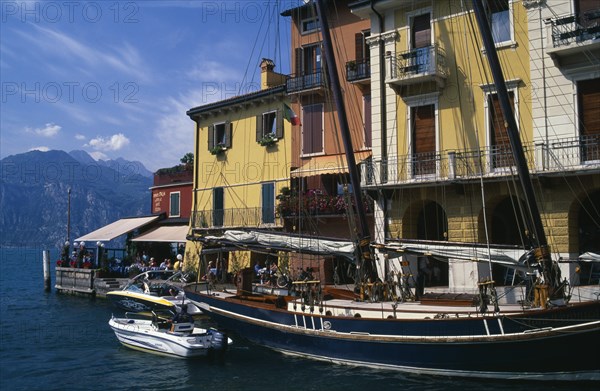 ITALY, Veneto, Malcesine, "Yellow, white and terracotta painted facades of waterside buildings with moored yacht and motor boats in foreground.  People at cafe at end looking out across lake "