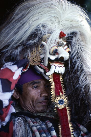 INDONESIA, Bali, Religion, Portrait of village man with head dress topped with mask depicting the witch Rangda who epitomises evil.  He becomes possessed by the spirit of Rangda during the performance of a ritual drama.