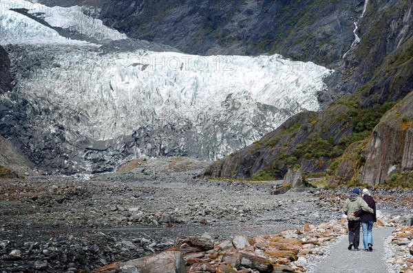 NEW ZEALAND, SOUTH ISLAND, WEST COAST, "MOUNT COOK NATIONAL PARK, A COUPLE WALK TO THE TERMINAL FACE OF THE FRANZ JOSEF GLACIER. Julius von Haast, geologist and explorer, named Franz Josef Glacier in 1863, after the Emperor of the Austro-Hungarian Empire. Approximately 7000 years old, and a remnant of a much older and larger glacier which originally swept right to the sea, Franz Josef Glacier extends 12 kilometres from its three feeder glaciers in the high snow fields of the southern Alps. "