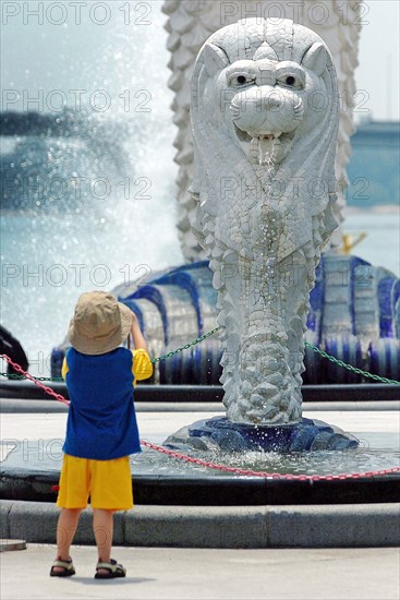 SINGAPORE, MARINA BAY, "A YOUNG BOY LOOKS ON AT A MINI VERSION OF THE MERLION STATUE. The Merlion is one of the most well-known tourist icons of Singapore. Its landmark statue, once at the Merlion Park, was relocated to the front of the Fullerton Hotel in April 2002.In 2002, the statue was relocated to its current site that fronts Marina Bay with the completion of the Esplanade Bridge in 1997. The statue measures 8.6 metres high and weighs 70 tonnes. The merlion was designed by Fraser Brunner for the Singapore Tourism Board in 1964 and was used as its logo up to 1997. The Merlion continues to be its trademark symbol."
