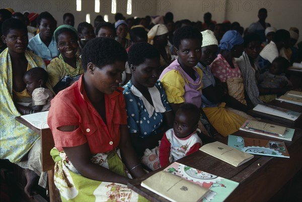 MALAWI, Adult Education, "Adult literacy class for refugees from Mozambique in Kunyinda Camp.  Classroom of young women, some with children sitting at long wooden desks."