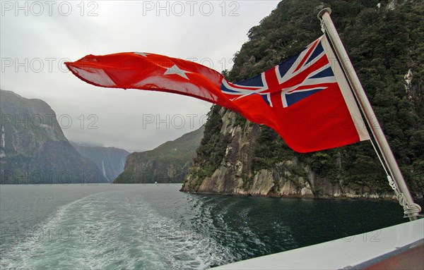 NEW ZEALAND, SOUTH ISLAND, "MILFORD SOUND,", "SOUTHLAND, A NEW ZEALND ENSIGN FLAG WAVES ON THE REAR OF REAL JOURNEYS CRUISE BOAT THE MILFORD MARINER AS IT TRAVELS ALONG MILFORD SOUND IN NEW ZEALANDS FJORDLAND AREA"