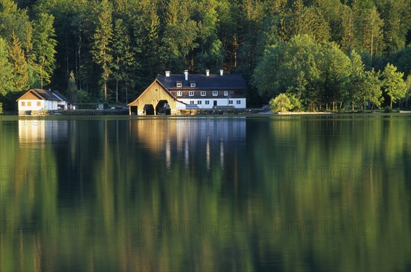 AUSTRIA, Oberosterreich, Mondsee, View across lake towards boat house and other building with forest backdrop reflected in flat surface of water.