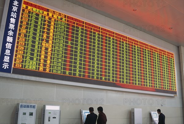 CHINA, Beijing, Interior of railway terminal with electronic information board with customers looking at smaller screens beside ticket machines below.