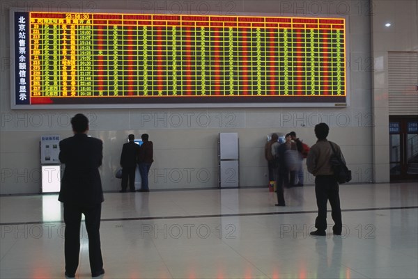 CHINA, Beijing, Interior of railway terminal with electronic information board with customers looking at smaller screens beside ticket machines below.