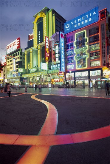CHINA, Shanghai, Nanjing Lu.  Busy street scene at night with illuminated neon signs and advertising and ribbon of pink / orange lit pavement across square in the foreground.