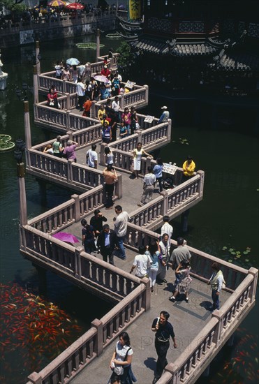 CHINA, Shanghai, "Yu Gardens.  View over zig-zag foot-bridge over pond with mass of silver and orange Koi carp. Vendors and crowds smoking, sightseeing and posing for / taking photograhs"