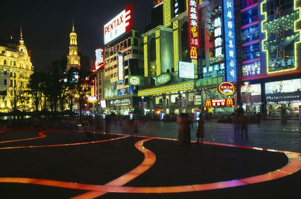 CHINA, Shanghai, "Nanjing Lu. Busy street scene at night with illuminated shop fronts, neon signs and advertising and ribbon of lit pink / orange paving across square in foreground."