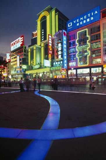 CHINA, Shanghai, Nanjing Lu.  Busy street scene at night with illuminated neon signs and advertising and ribbon of blue lit pavement across square in the foreground.