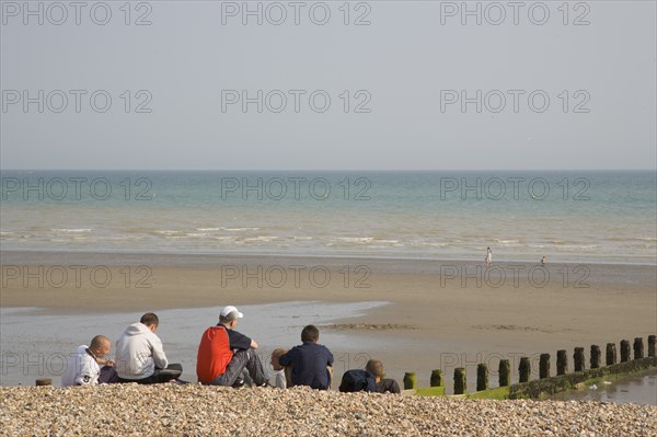 ENGLAND, West Sussex, Bognor Regis, A group of teenage boys sitting on shingle and sand beach looking out towards the sea