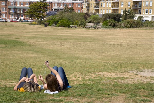 ENGLAND, West Sussex, Littlehampton, Two girls lying on grass by seafront promenade using and sharing an MP3 music player