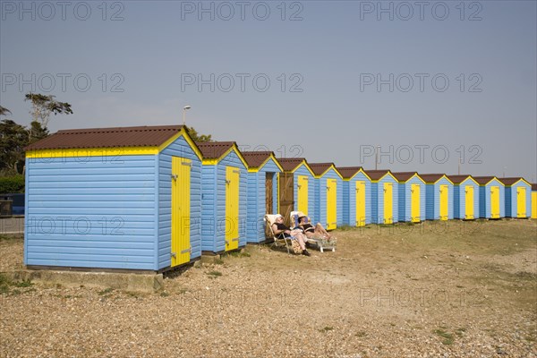ENGLAND, West Sussex, Littlehampton, A crescent of blue and yellow beach huts with a couple sunbathing on loungers