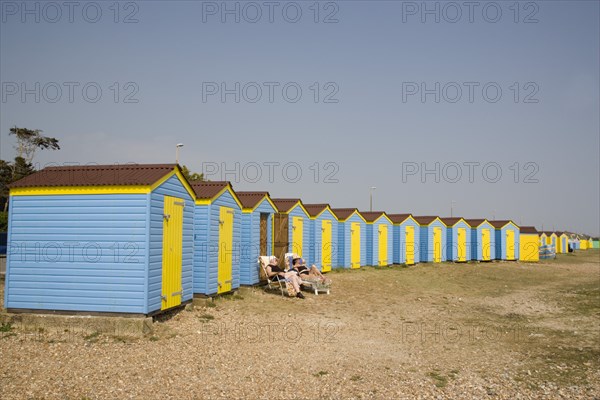 ENGLAND, West Sussex, Littlehampton, A crescent of blue and yellow beach huts with a couple sunbathing on loungers