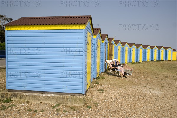 ENGLAND, West Sussex, Littlehampton, A crescent of blue and yellow beach huts with a couple sunbathing on loungers