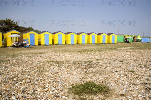 ENGLAND, West Sussex, Littlehampton, A crescent of  yellow and blue beach huts with sunbathers on loungers