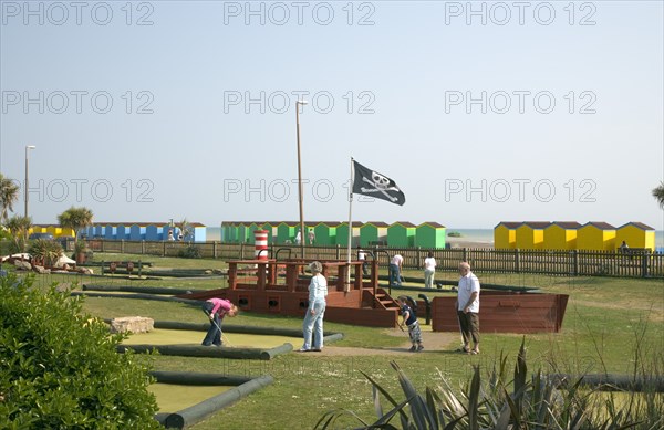 ENGLAND, West Sussex, Littlehampton, Families enjoying pitch and putt mini golf games in Norfolk Gardens next to seafront. A skull and cross bones flag flying. Colourful beach huts behind.