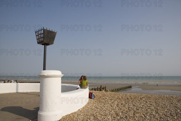 ENGLAND, West Sussex, Bognor Regis, Two young women sitting on a white wall facing each other talking near a torch beacon. Sand and shingle beach with families seen at the waters edge behind