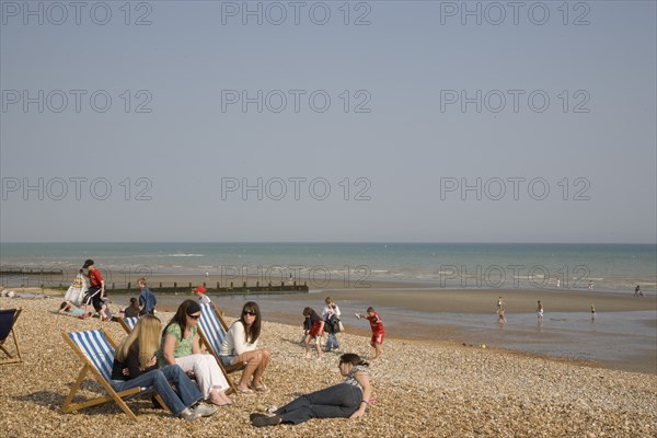 ENGLAND, West Sussex, Bognor Regis, A group of girls sitting on deck chairs on shingle beach with children playing behind