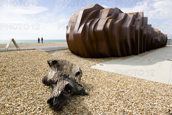 ENGLAND, West Sussex, Littlehampton, Two people standing on the beach looking at the rusted metal structure of the fish and seafood restaurant the East Beach Cafe designed by Thomas Heatherwick with an A-frame on the promenade and driftwood on the pebble beach in the foreground