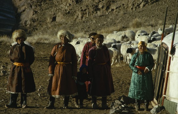 MONGOLIA, Gobi Desert, Biger Negdel, Khalkha winter sheep camp with complete family  father mother daughter grandfather and grandchildren standing outside family ger yurt with sheep in stone walled pen and mountainside in background