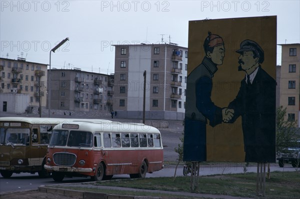 MONGOLIA, Ulan Bator, Soviet billboard in central Ulan Bator  depicting Lenin shaking hands with Mongolia's first president Sukhebator in urban area with multi-storey apartment blocks behind and buses moving along street. Ulaanbaatar East Asia Soviet Mongolia Asian Baator Male Men Guy Mongol Uls Ulaan