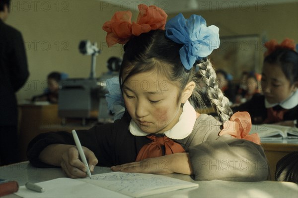 MONGOLIA, Education, Altai SecondarySchool  11 year old girl writing at desk with her hair plaited and tied with blue and orange bows. East Asia Asian Kids Learning Lessons Mongol Uls Mongolian Teaching