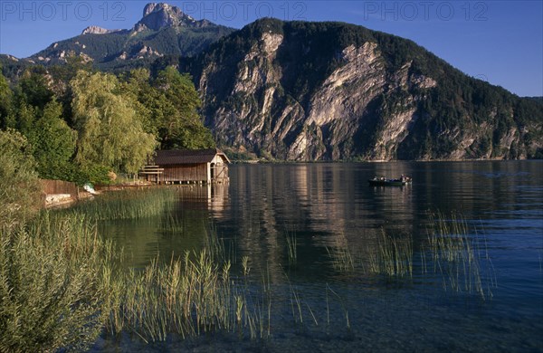 AUSTRIA, Oberosterreich, Mondsee, "Small motor boat approaching boat house on shore of Monsee Lake with mountain backdrop reflected in rippled surface, reeds and willow trees."
