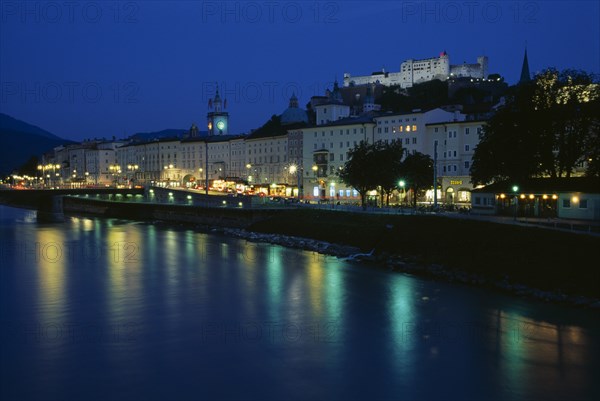 AUSTRIA, Salzburg, City view at night with green and yellow lights of river side street lamps and buildings reflected on water surface. Hohensalzburg Fortress elevated on hillside behind.