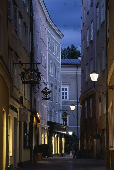 AUSTRIA, Salzburg, "Narrow street of Judengasse at night lined by tall, grey, pink and cream painted buildings with street lamps extending from walls on metal brackets."